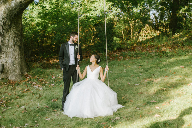 groom looks at bride who is seated on a tree swing