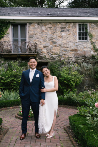 bride and groom pose in a garden in front of a brick building