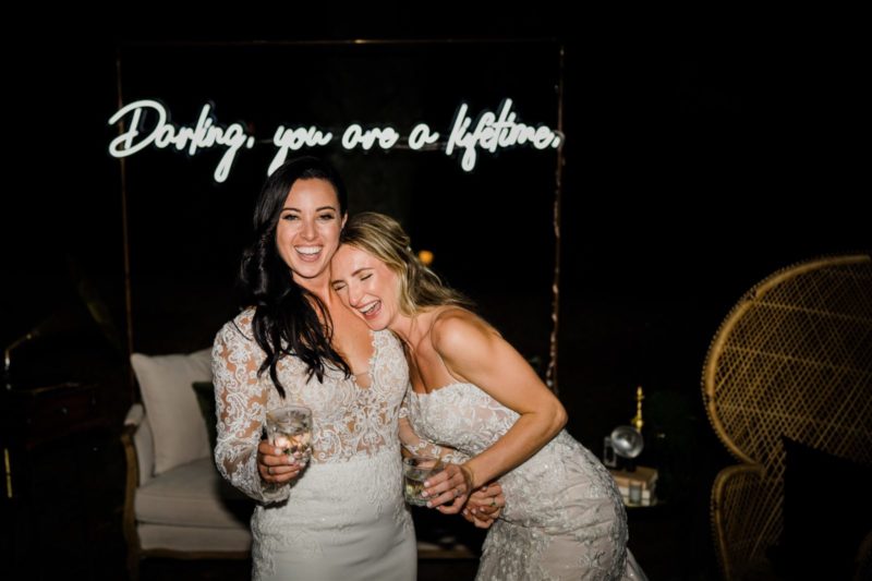 two brides smiling together in front of a neon sign