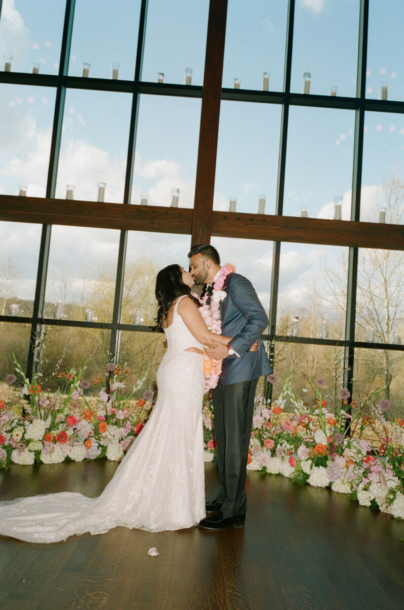 Just married kiss, farmhouse ceremony with Bright pink, orange, and purple floral horseshoe arbor for Indian wedding ceremony with carnation and rose garlands