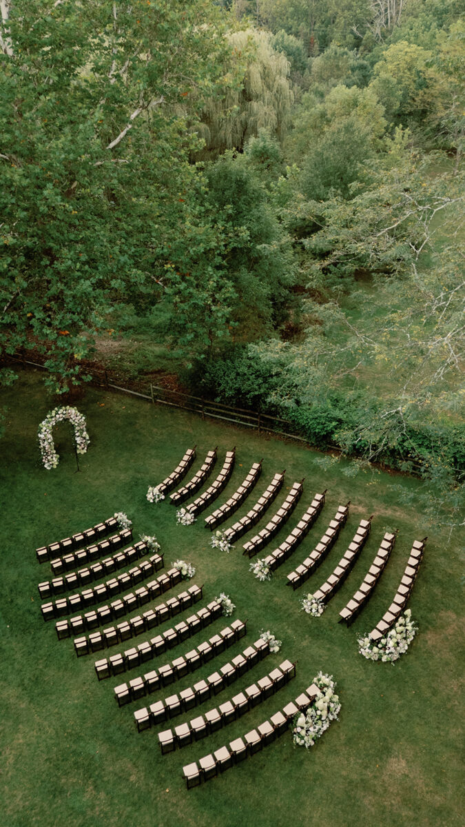 Drone view of ceremony under oak trees at New Jersey estate wedding. Aisle lined with dark fruitwood chairs and white blush and green floral arrangements. September garden wedding