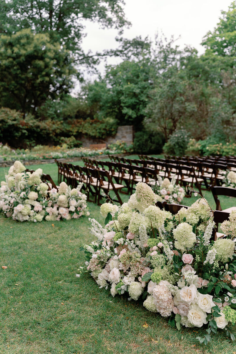 Aisle lined with dark fruitwood chairs and white blush and green floral arrangements