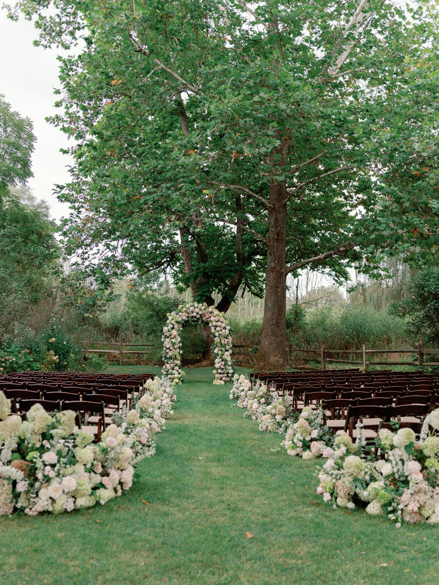 Organic floral ceremony arbor with hydrangeas and roses under oak trees. Aisle lined with dark fruitwood chairs and white blush and green floral arrangements