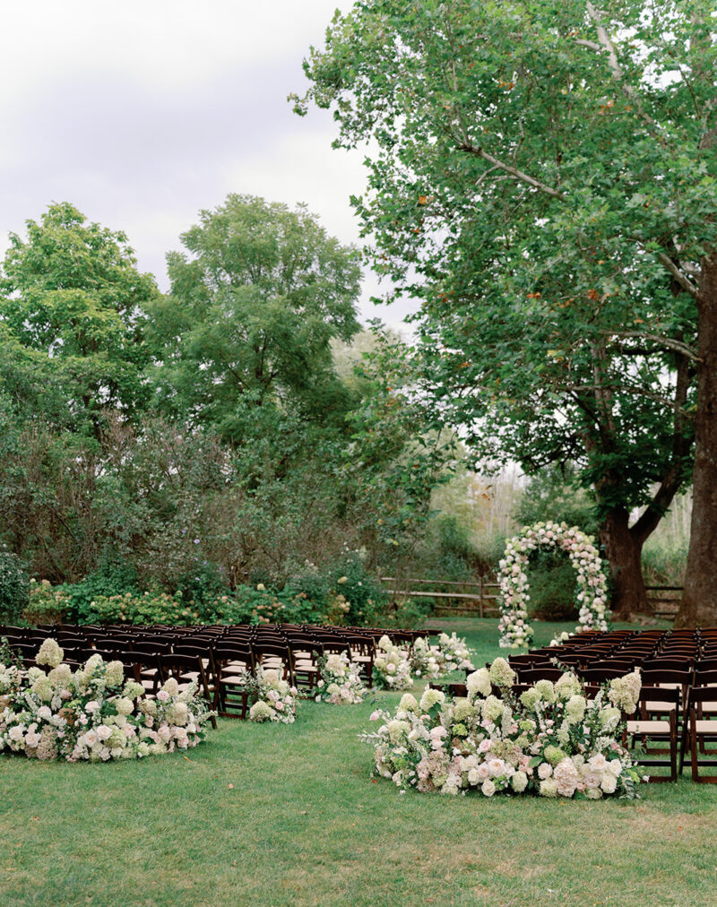 Aisle lined with dark fruitwood chairs and white blush and green floral arrangements